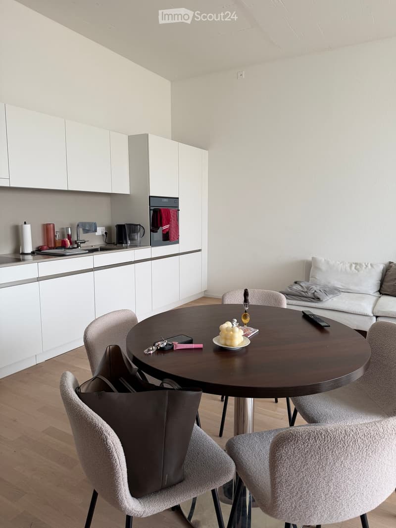 A modern, open-plan living and dining area with a round wooden dining table, four chairs, a sofa, and a kitchen area with white cabinets, a sink, and an oven.