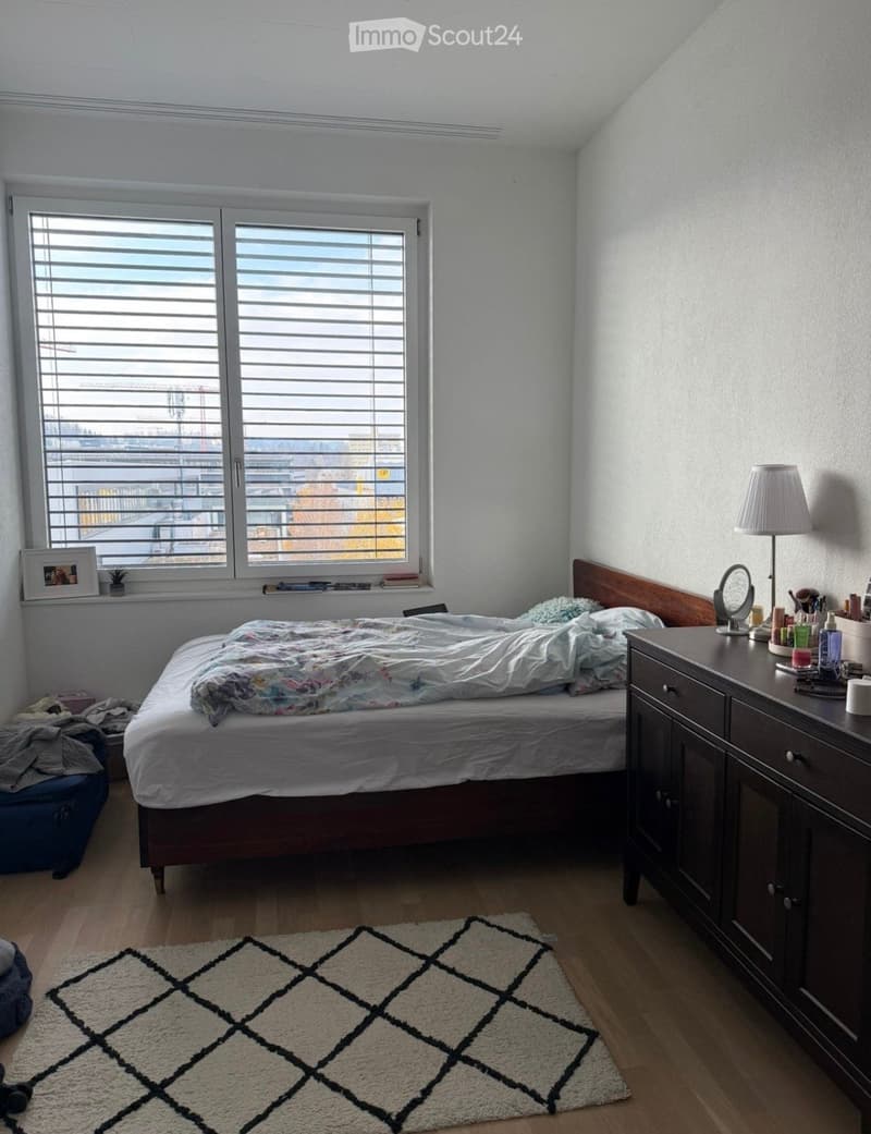 A cozy bedroom with a wooden bed, a nightstand with a lamp, a mirror, and a patterned rug on the floor. The room features large windows with blinds and a view of the city.