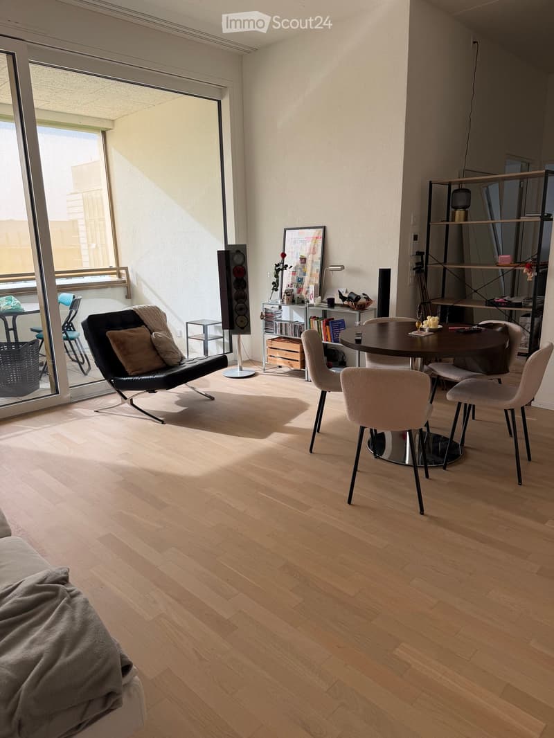 Modern living room with wooden flooring, large windows, balcony access, sofa, chairs, round table, shelves, and speakers.
