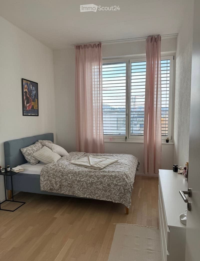 Modern bedroom with wooden flooring, large window with pink curtains, bed with patterned bedding, nightstand, and a small table with items.
