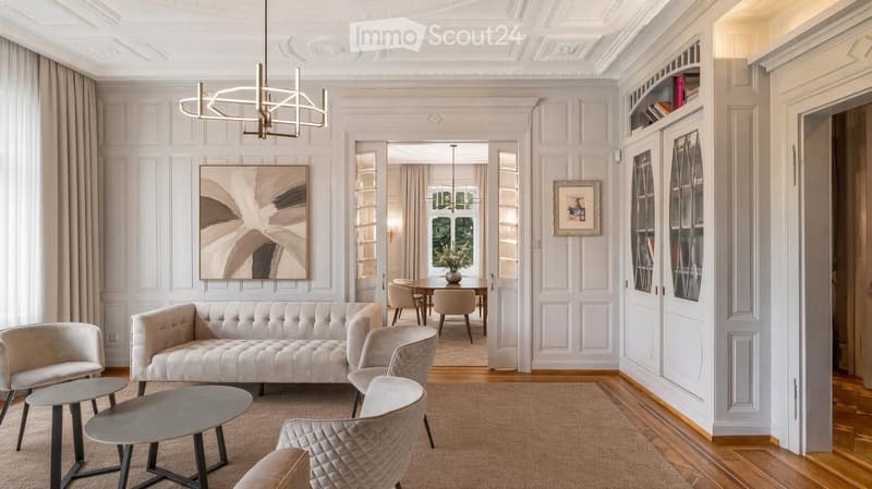 Elegant living room with a cream-colored sofa, coffee table, armchairs, dining area, bookshelf, and artwork.