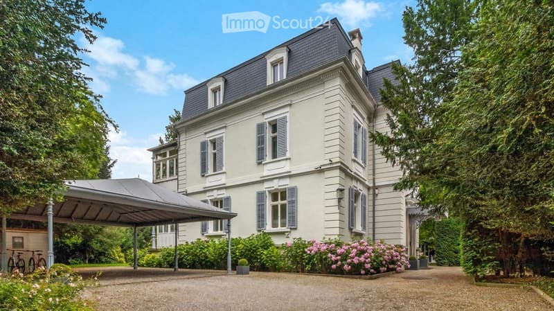 A large, elegant two-story house with light beige walls, dark grey roof, multiple windows with shutters, and a covered driveway. The house is surrounded by lush greenery and flowering bushes.