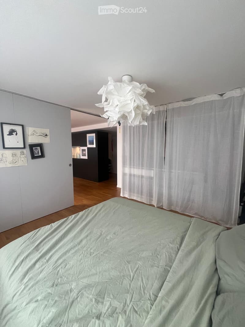 A modern bedroom with a neatly made bed, white curtains, and several picture frames on the wall. A hallway leads to another room with dark walls and additional artwork.