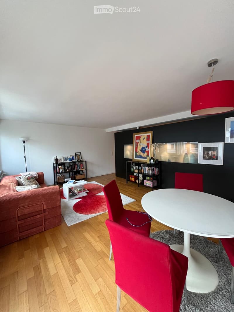 Modern living room with wooden flooring, red sofa, white dining table with red chairs, bookshelf, and decorative items.