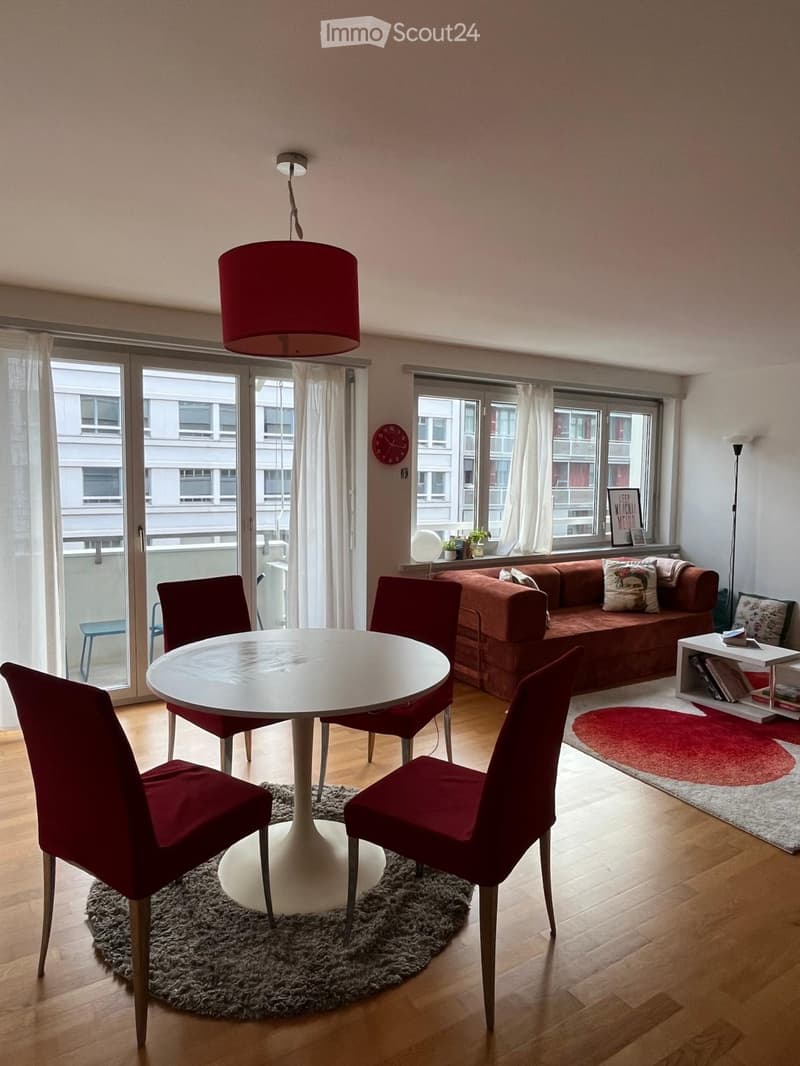 Modern living room with wooden flooring, large windows, a red sofa, a white round table with four red chairs, a hanging lamp, and a side table with books and a picture frame.