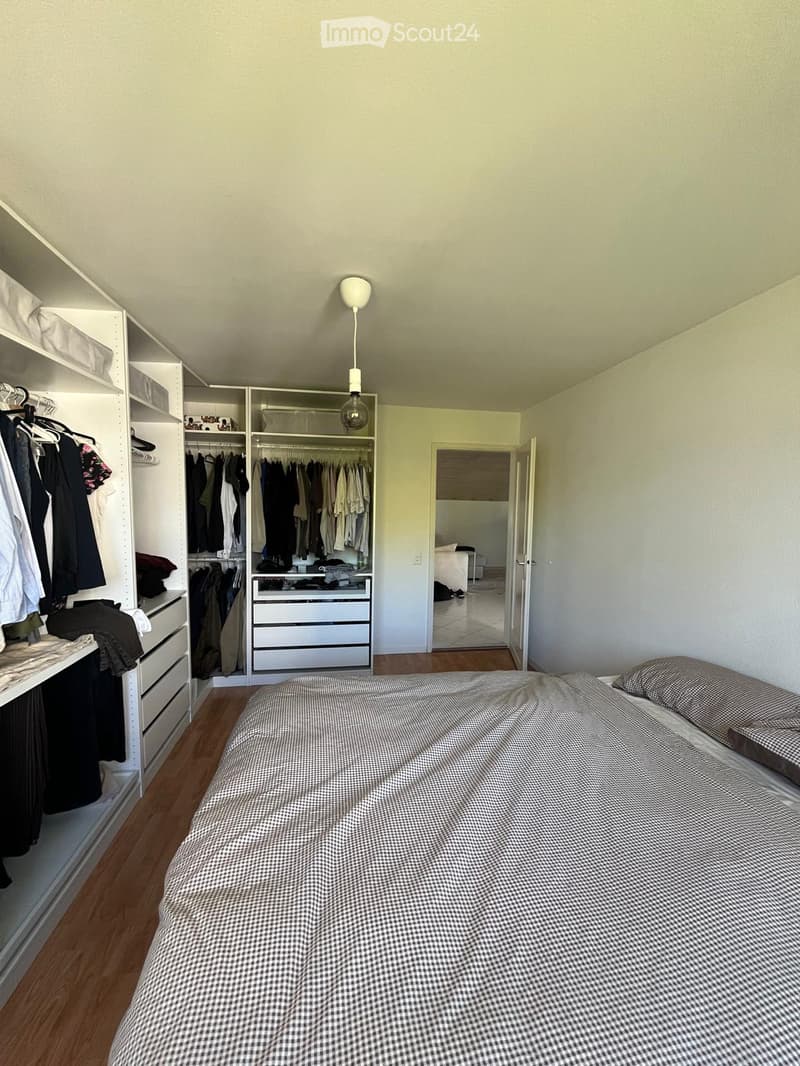 A modern bedroom with a neatly made bed, a large built-in wardrobe with multiple sections, and a set of drawers. The room features wooden flooring and a hanging light fixture.