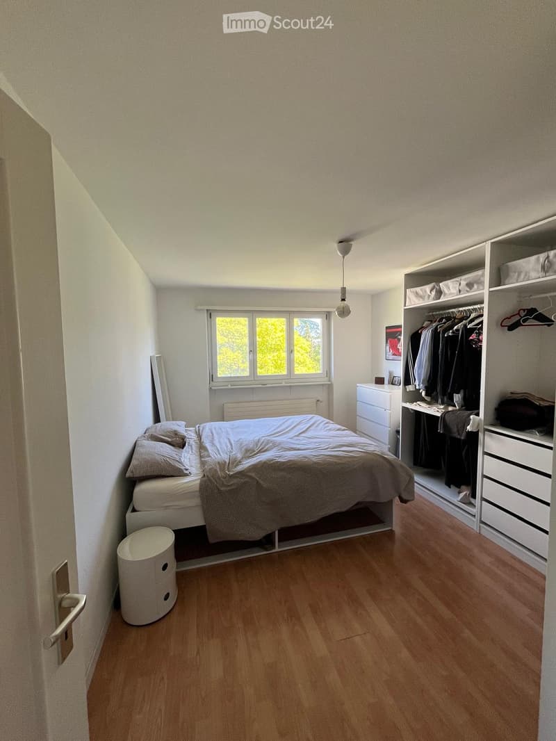 Cozy bedroom with wooden flooring, a bed with pillows and a blanket, a wardrobe with clothes, a small drawer, and a round stool near the door.