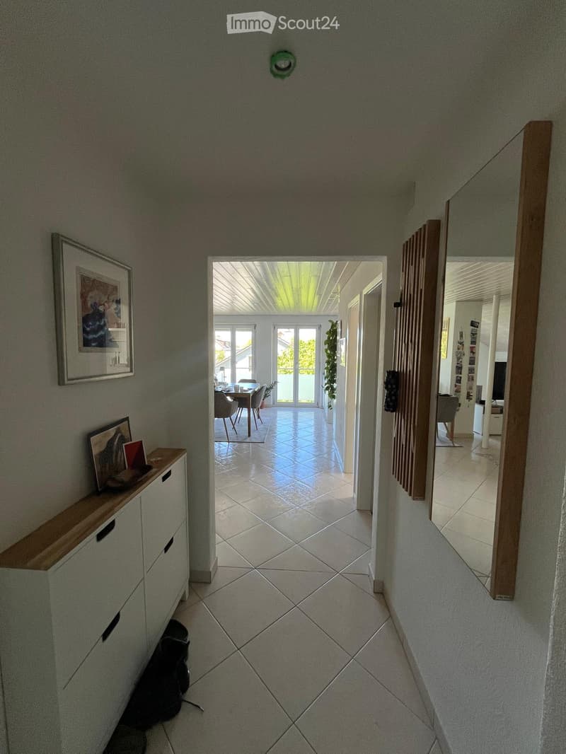 A well-lit hallway with tiled flooring, leading to a dining area and other rooms. Features include a small wooden shelf with framed pictures, a mirror, and a cat sitting on the floor.