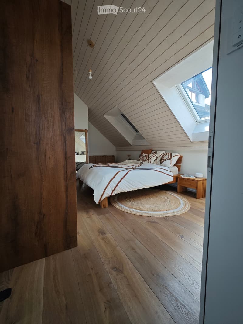 Cozy attic bedroom with wooden floors, slanted wooden ceiling, skylight windows, and a neatly made bed with pillows and a duvet.