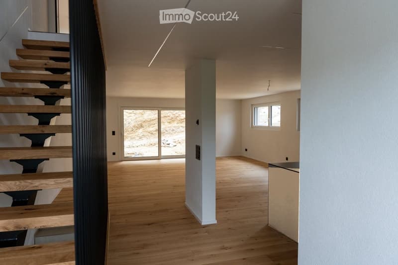 Empty room with wooden flooring, white walls, and a staircase leading upwards. The room has large windows and a door leading to an outdoor area.