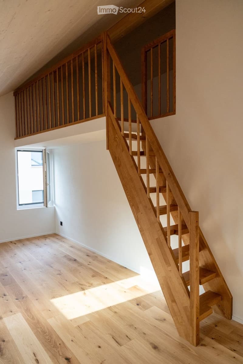Wooden staircase with a modern design, light wooden flooring, and white walls. The staircase features a wooden railing and steps, with a small window allowing natural light to enter the space.