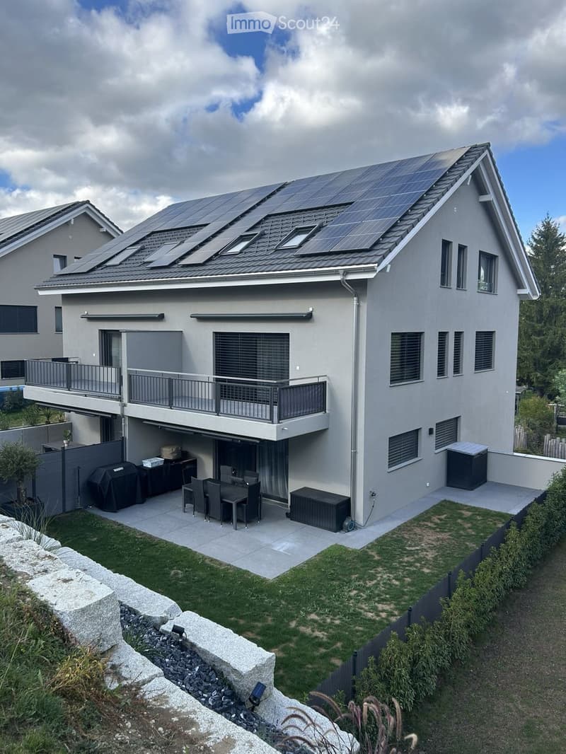 Modern two-story house with solar panels on the roof, large windows, balcony, garage, and outdoor patio with dining set and grill.