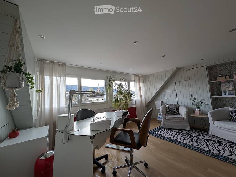 Bright, modern room with wooden flooring, large windows, a desk with office chair, a sofa, a small table, and decorative plants.