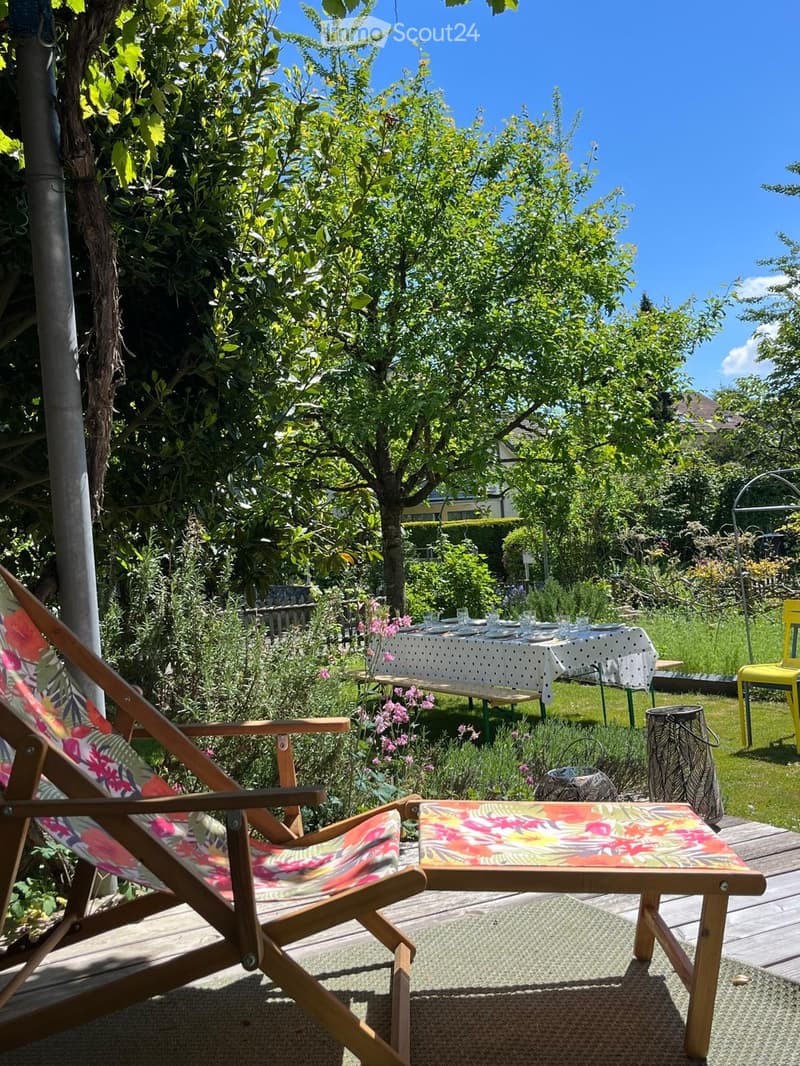 Garden with a wooden deck, a table with a white tablecloth, chairs, a bench, and a small stool, surrounded by greenery and flowers.