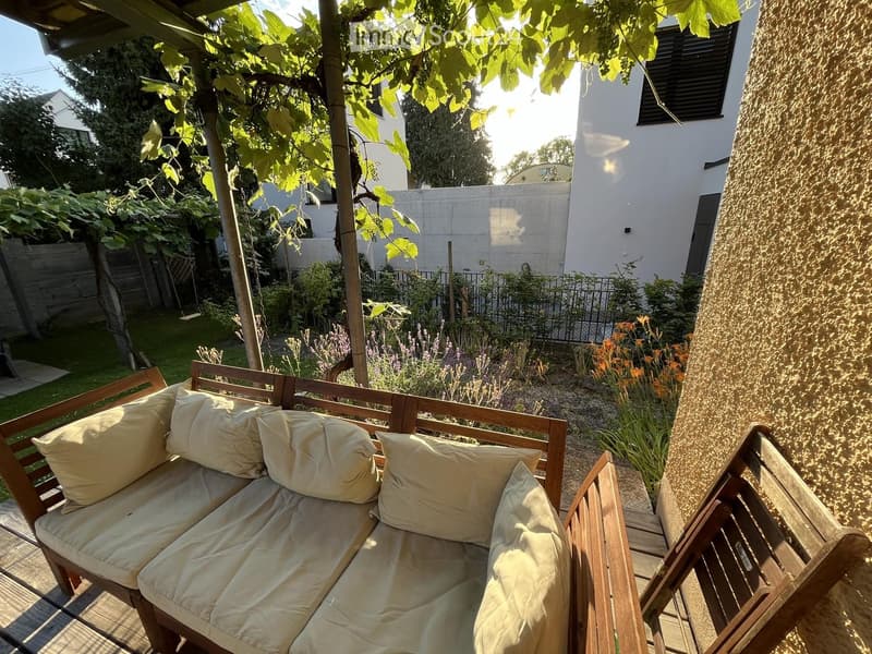 Wooden balcony with a cream-colored sofa and pillows, surrounded by greenery and a garden area.