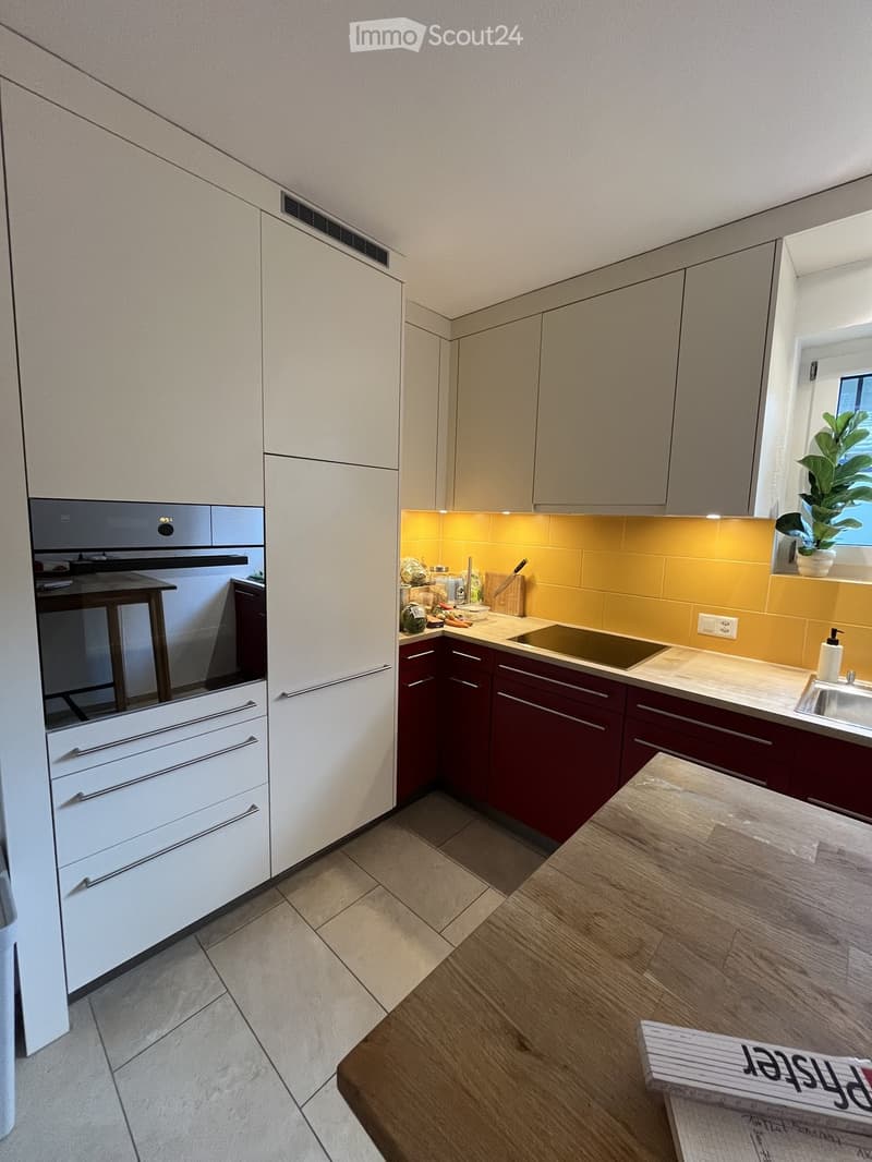 Modern kitchen with white and dark red cabinets, a central island, and a built-in stove. Features include a sink, a wooden table, and a potted plant on the windowsill.