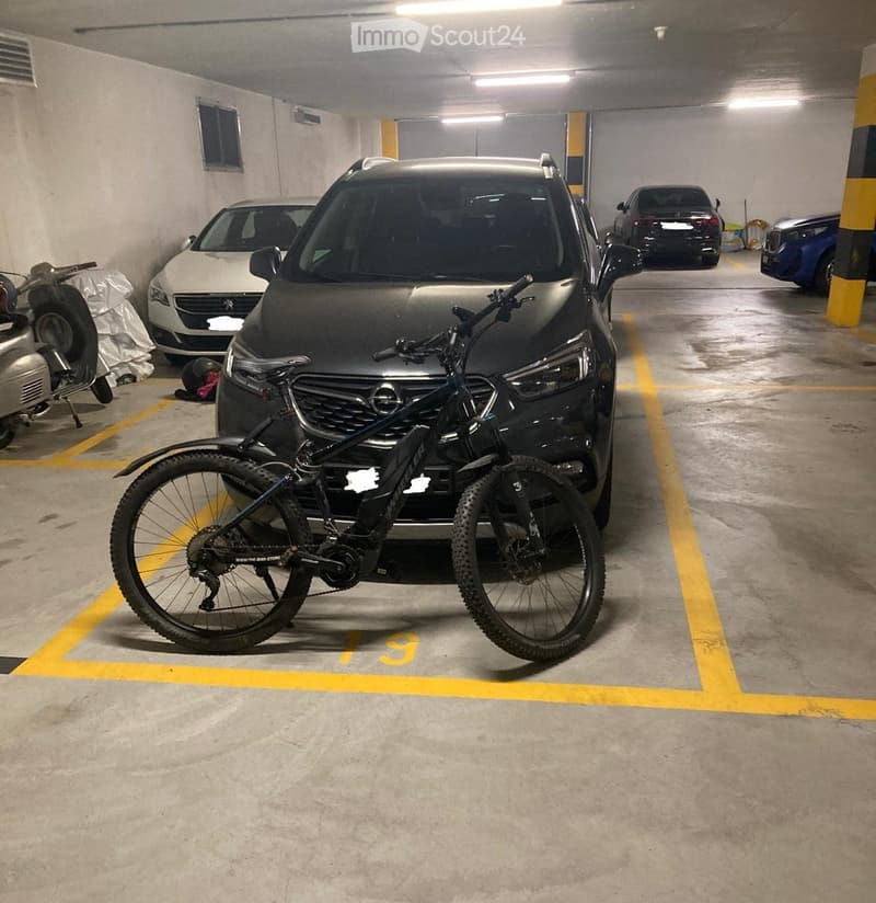 Underground parking area with several parked cars, a bicycle, and a scooter. The area is marked with yellow lines and features concrete floors and walls.