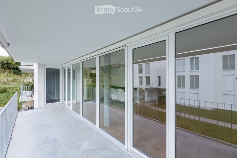 Modern balcony with large glass sliding doors, white metal railing, and a view of neighboring buildings and greenery.