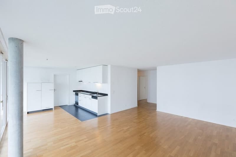Empty room with wooden flooring, white walls, and a small kitchen area featuring white cabinets and a black countertop.