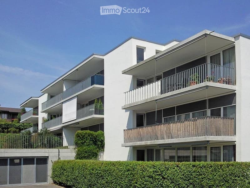 Modern multi-story apartment building with white exterior, multiple balconies, and greenery around the base.