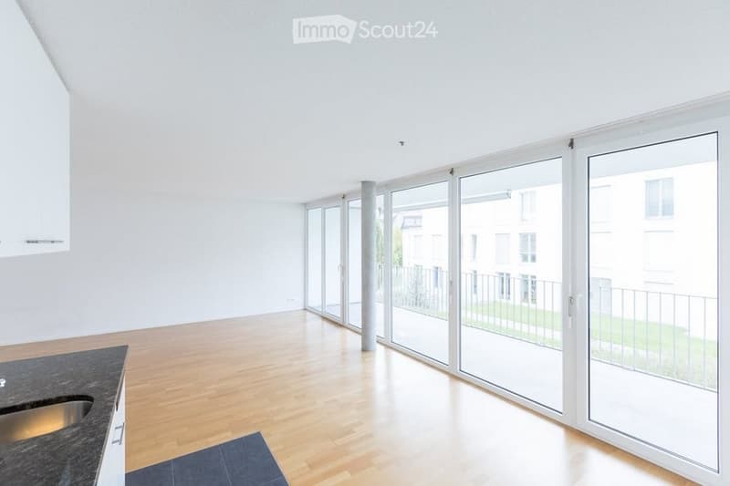 Bright, empty room with wooden flooring, large glass sliding doors leading to a balcony, and a small kitchen area with a sink.