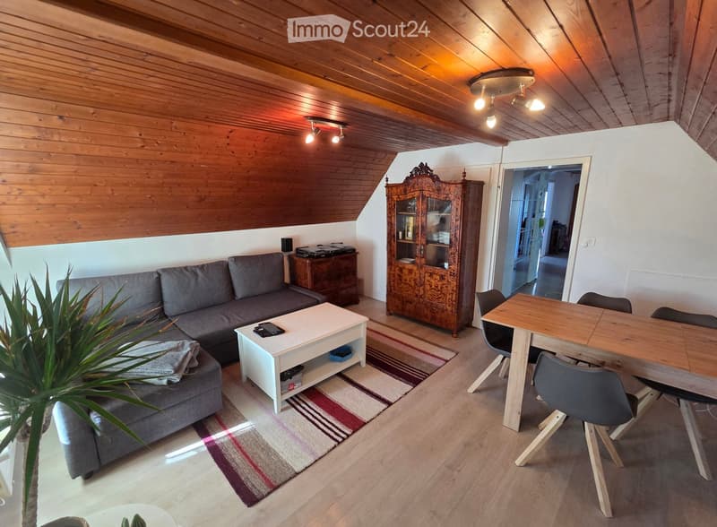 A modern living room with a grey sofa, white coffee table, wooden dining table with chairs, and a decorative cabinet. The room features wooden flooring, exposed wooden ceiling beams, and warm lighting.
