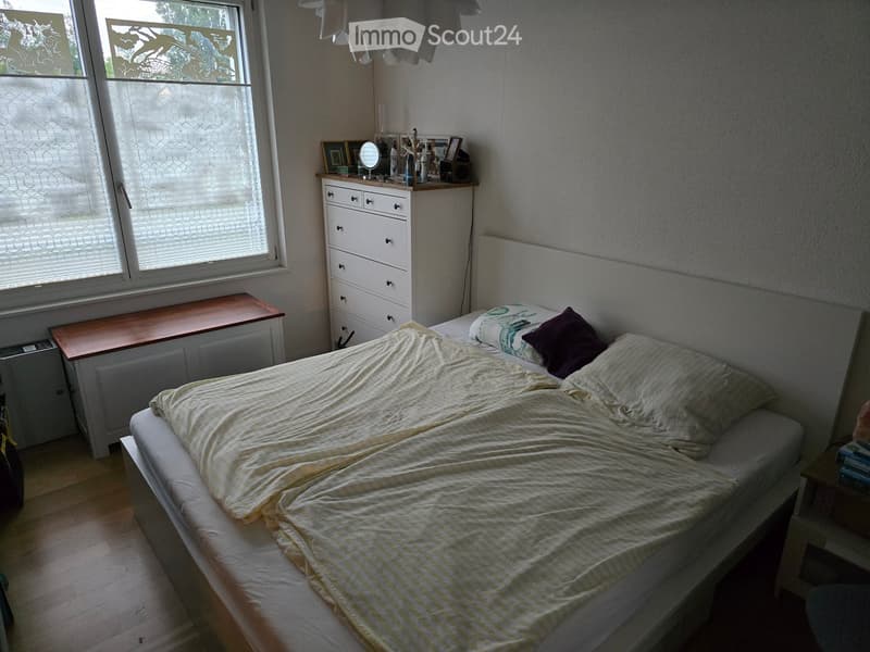Bedroom with a made bed, nightstand, mirror, drawer unit, and a small table near the window.