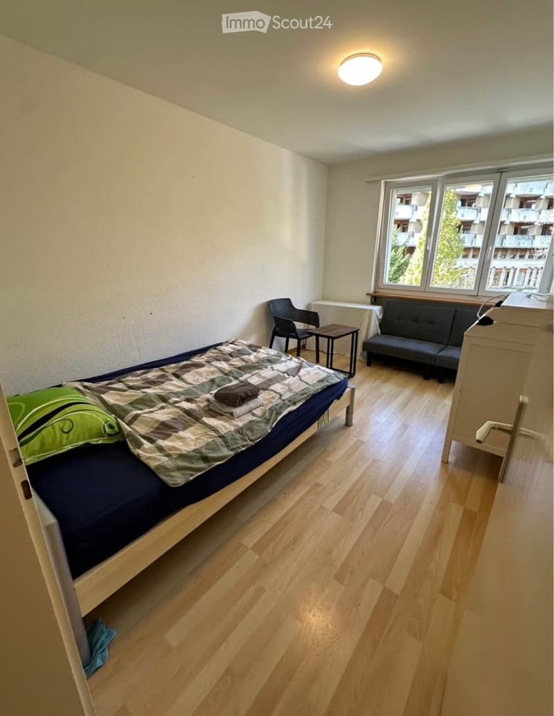 A small, furnished room with a single bed, a sofa, a chair, a small table, and a nightstand. The room has wooden flooring and large windows with a view of the outside.
