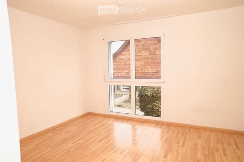 Empty room with light-colored walls and wooden flooring, featuring large windows and a sliding glass door leading to an outdoor area.