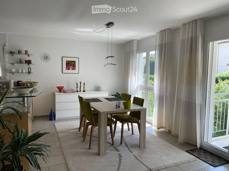 Modern dining area with a light wood dining table and four green chairs, a kitchen island with shelves, and large windows with curtains. A framed picture hangs on the wall, and a potted plant adds a touch of greenery.