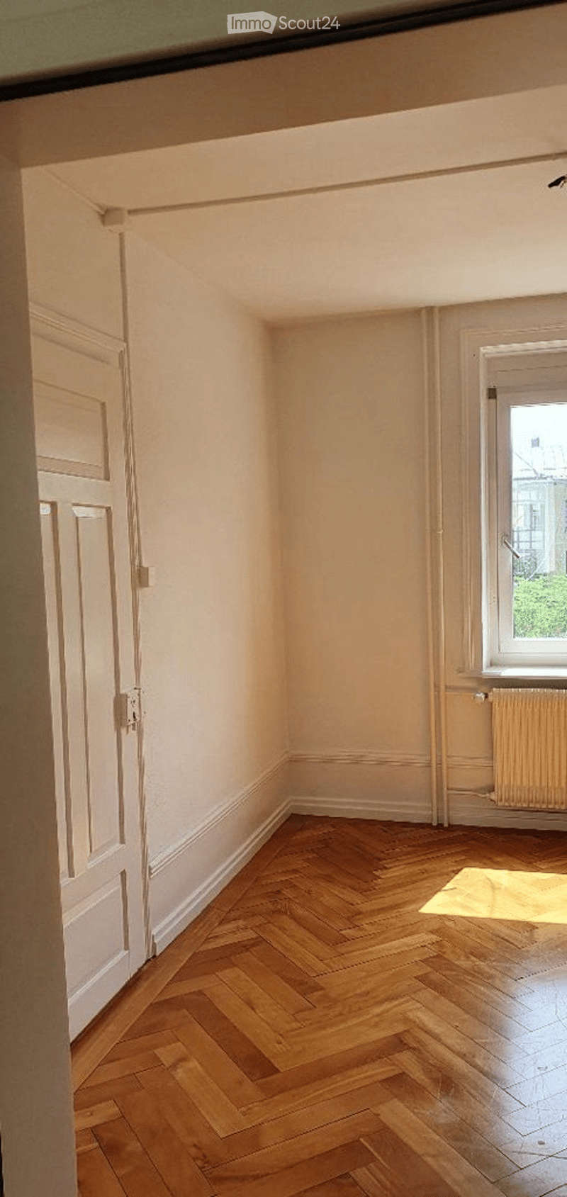 Empty room with wooden herringbone flooring, white walls, and a single window with a radiator below it. The room has a door on one side and is well-lit with natural light.