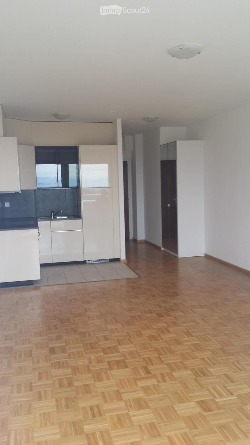 Empty room with wooden flooring and white walls, featuring a modern kitchen area with white cabinets and stainless steel appliances.