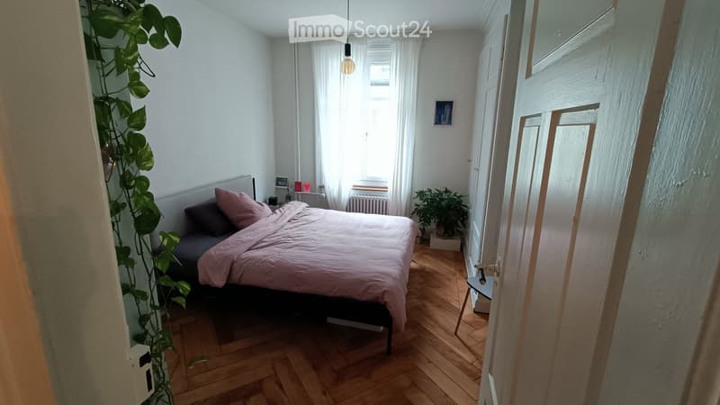 Cozy bedroom with wooden flooring, a bed with pink bedding, a nightstand, a small stool, and a potted plant near the window.