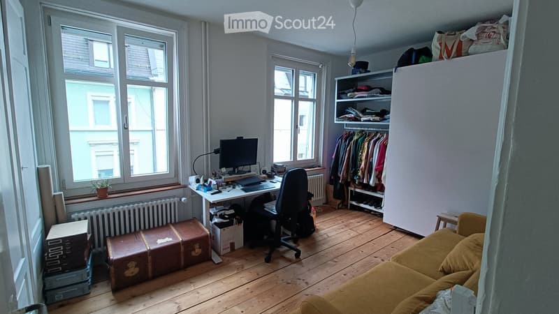 A furnished room with wooden flooring, large windows, a sofa, a desk with a laptop and monitor, a chair, a wardrobe, and a shelf with various items.