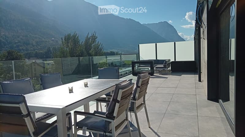 Modern outdoor terrace with a panoramic mountain view, featuring a white dining table with chairs, a sofa, and a glass railing.