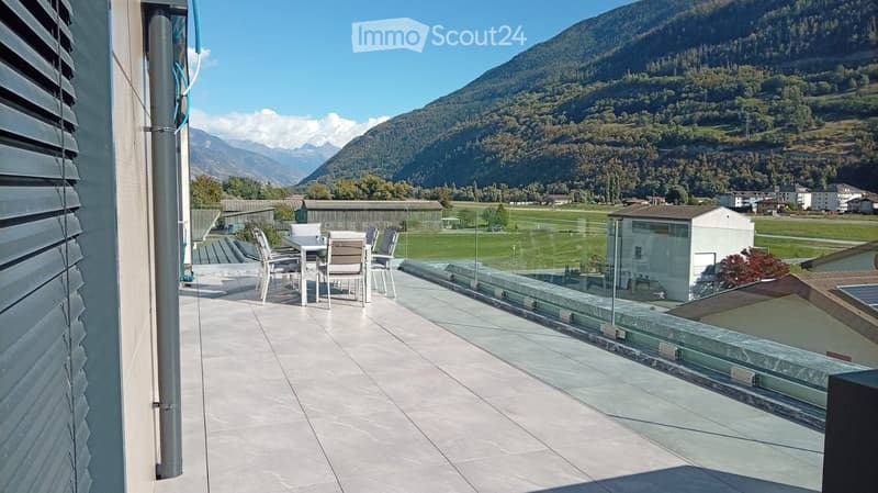 Balcony with a table and chairs, overlooking a scenic mountain landscape and green fields.