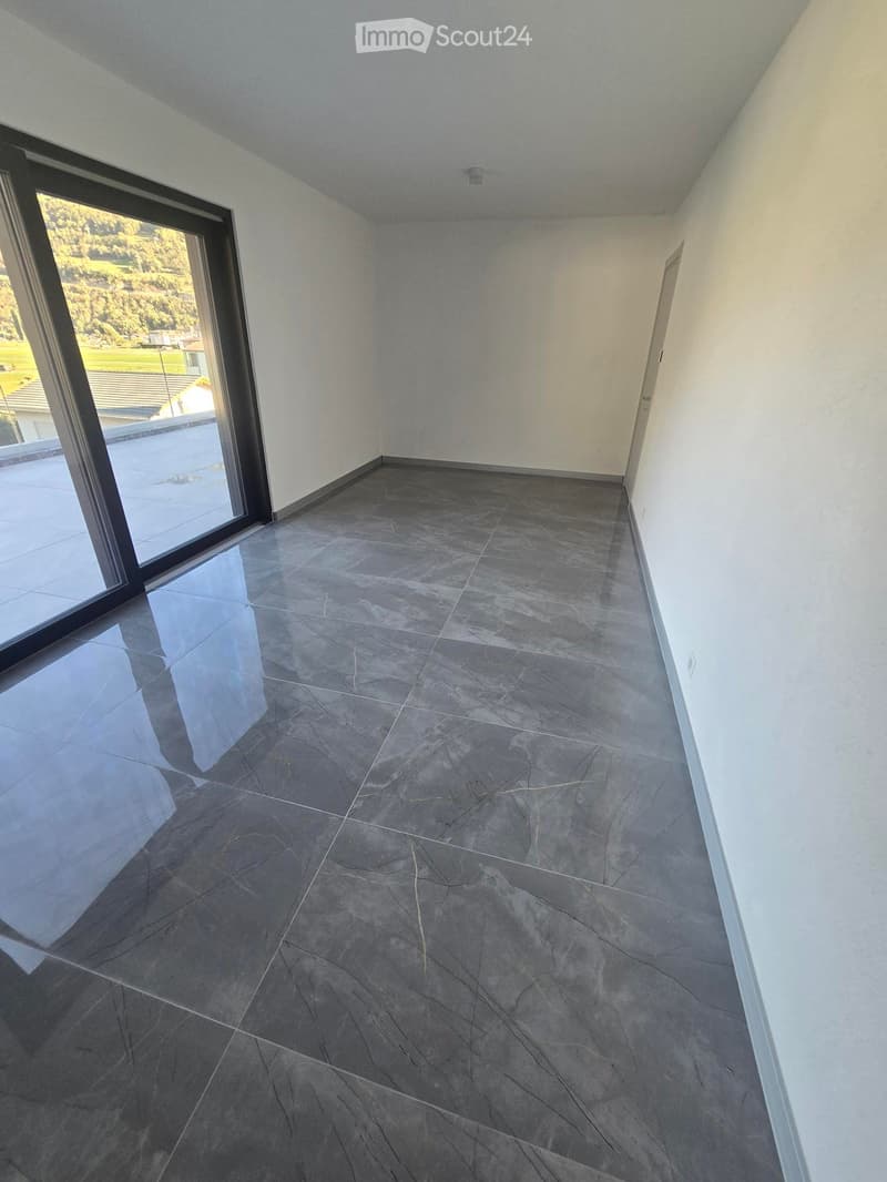 Empty room with grey tiled floor, white walls, large window with view of outdoor area, and a door leading to another space.