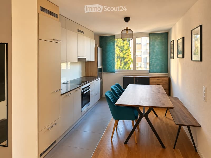Modern kitchen with white cabinets, stainless steel appliances, and a dining area featuring a wooden table, bench, and teal chairs.