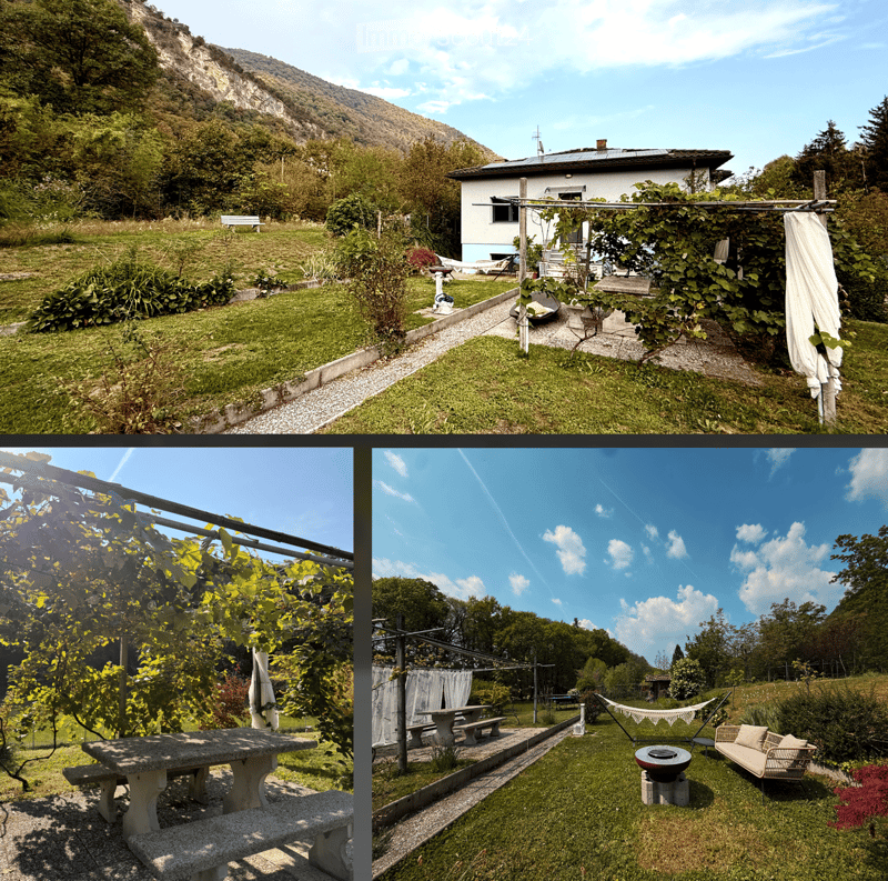 A scenic outdoor area with a house, garden, patio, and surrounding greenery. Features include a stone table, benches, a hammock, and a fire pit, with a mountain view in the background.