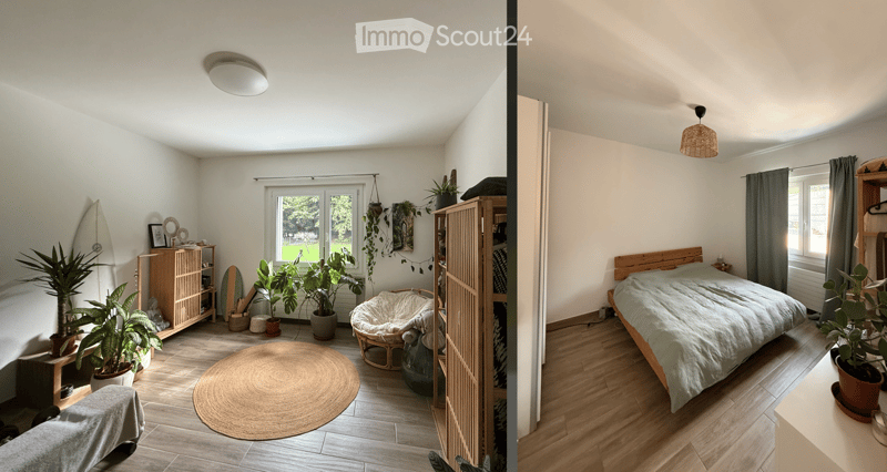 Cozy bedroom with a wooden bed, wicker chair, round jute rug, potted plants, and a surfboard leaning against the wall. The room features light wooden flooring and large windows with grey curtains.