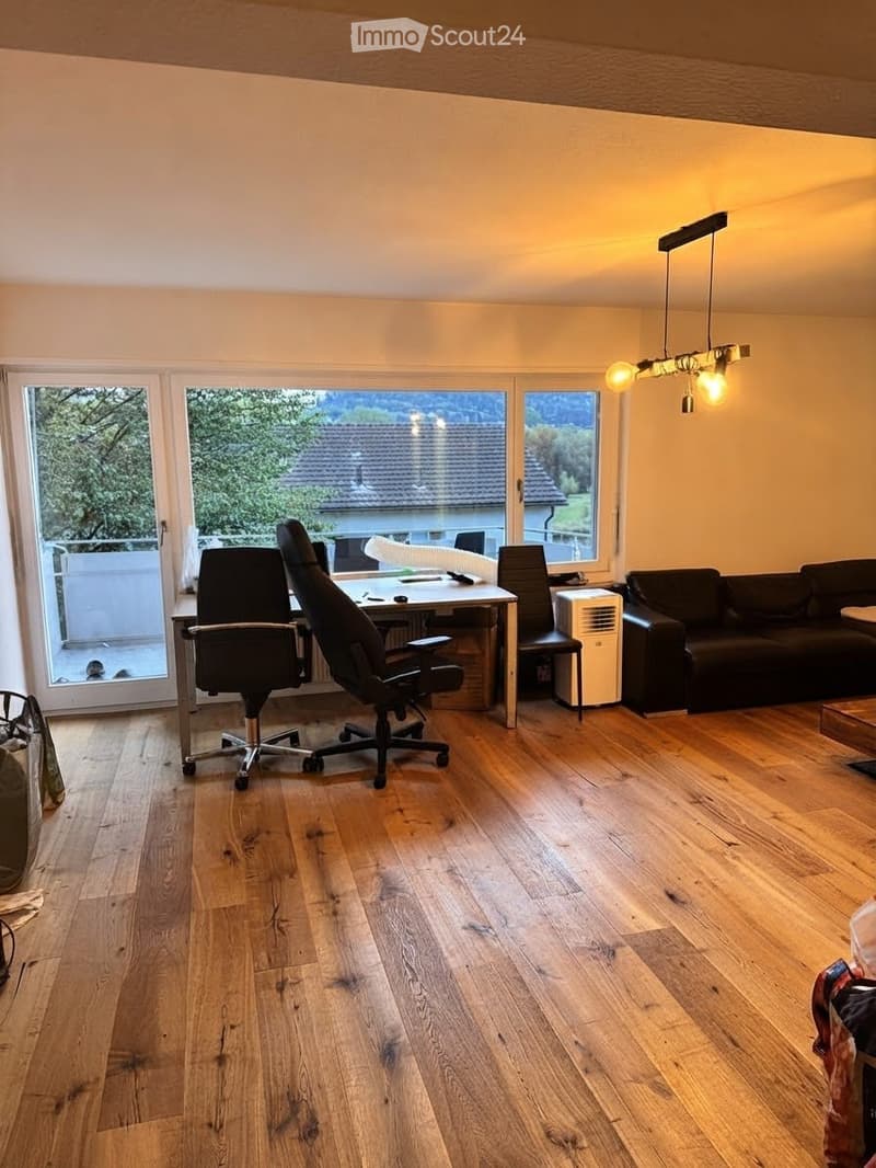 Bright living room with wooden flooring, large windows, a sofa, office chair, desk with laptop, and an air conditioner unit.