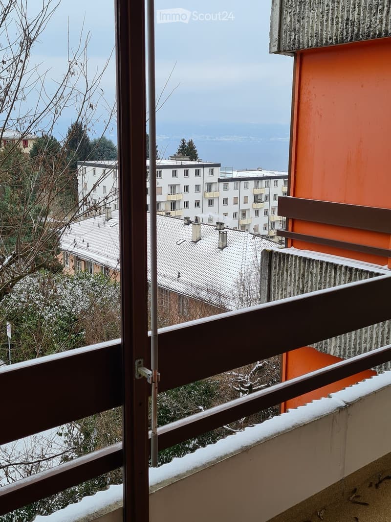 Balcony with wooden railing, overlooking a snowy urban area with buildings and distant water.