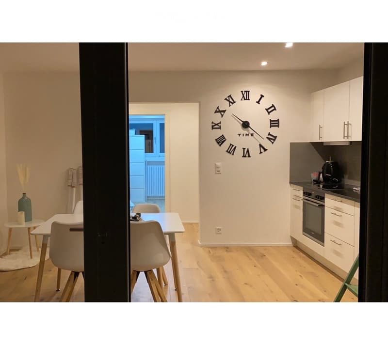 A modern, open-plan living and dining area with a kitchen. The space features a large wall clock, white dining table with chairs, a small side table with a vase, and a kitchen area with cabinets, an oven, and a coffee maker.