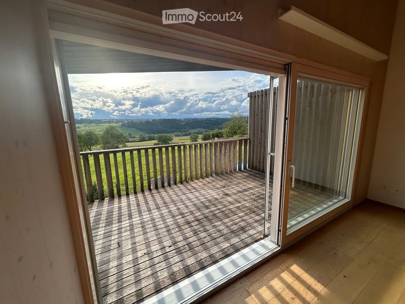 Open sliding glass door leads to a wooden balcony with a railing, overlooking a green field and distant landscape.