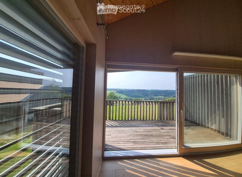 Bright, unfurnished room with large windows and sliding doors leading to a wooden balcony, offering a scenic view of the countryside.
