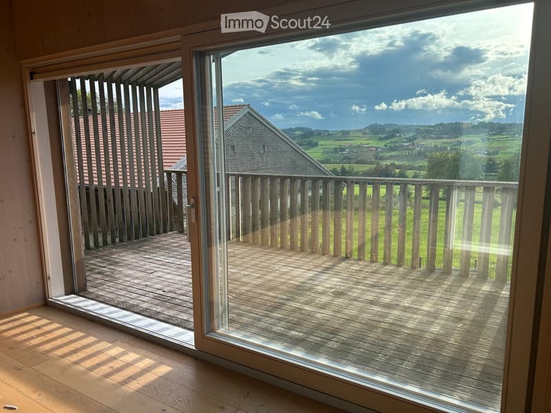 Open sliding glass doors lead to a wooden deck with a railing, overlooking a scenic countryside with fields and a distant house.