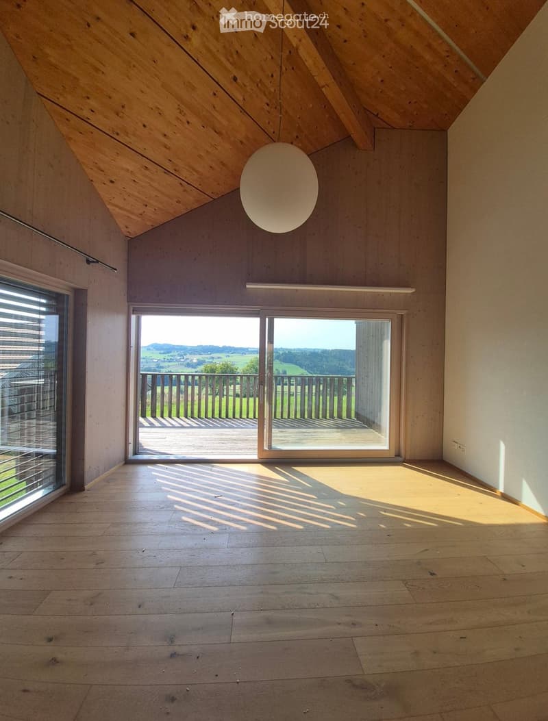 Bright, empty room with wooden flooring and slanted wooden ceiling. Large windows and sliding doors open to a balcony with a scenic view.