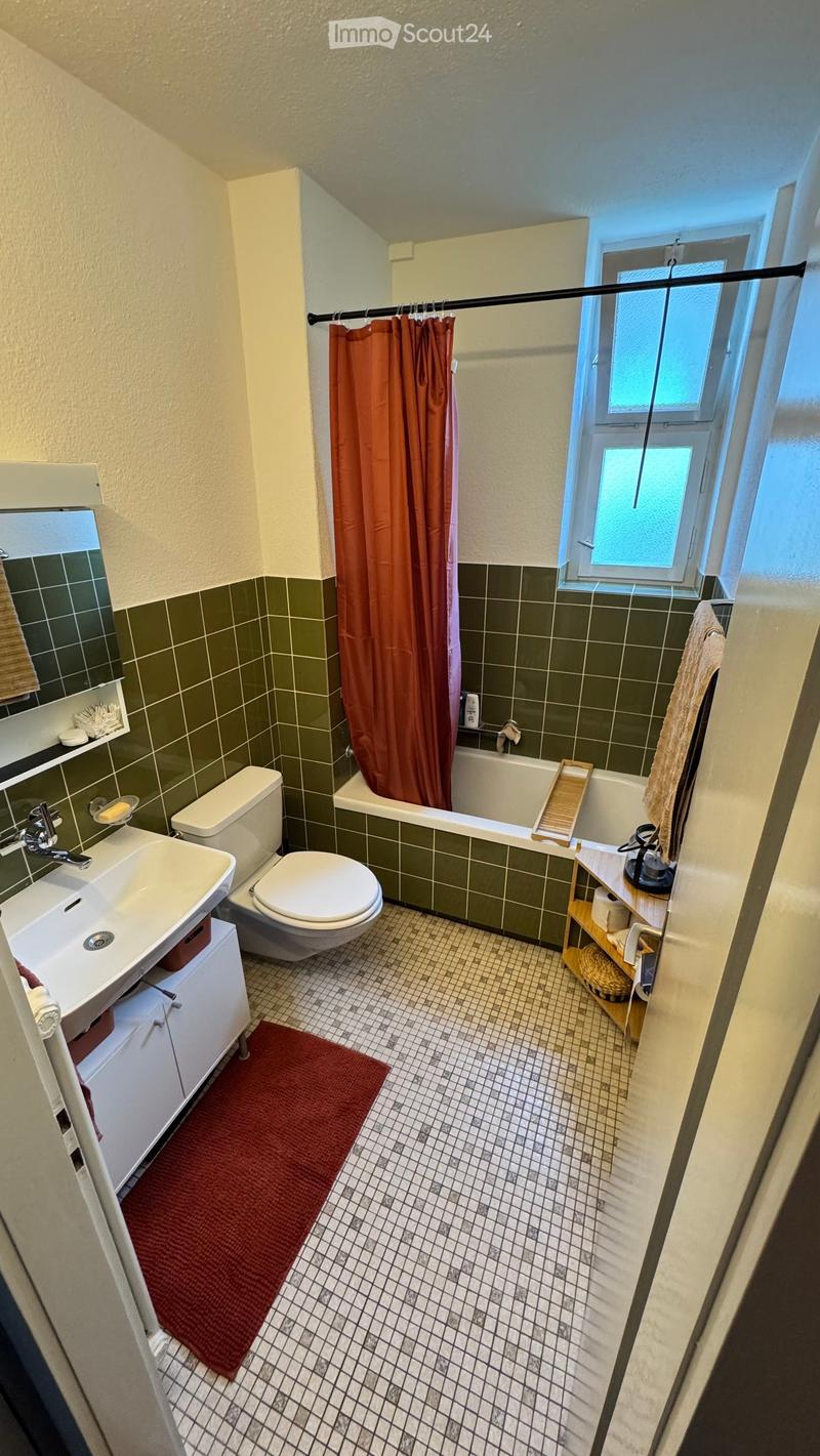 Bathroom with green tiled walls, white fixtures, a red bathmat, and a window with a curtain.
