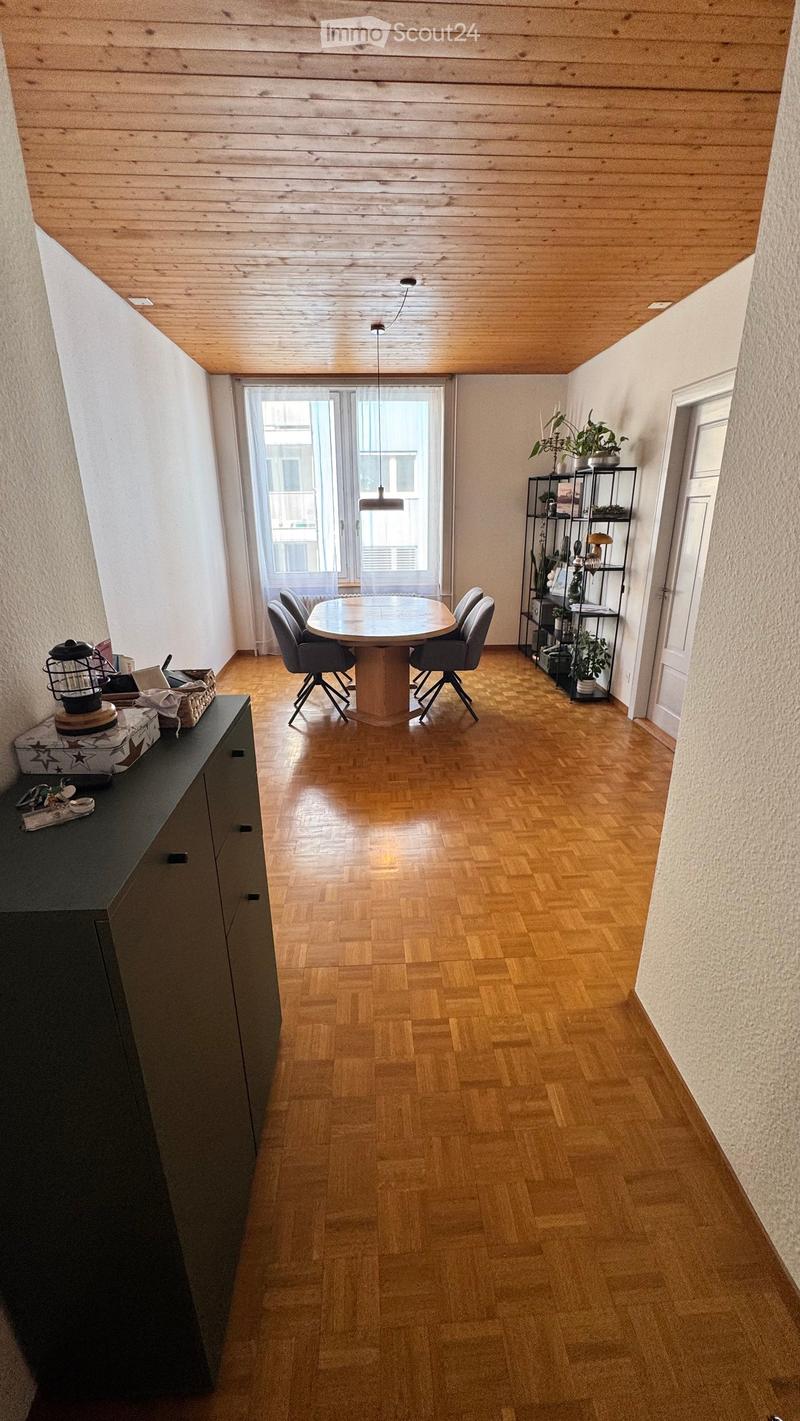 Dining area with wooden floor, round table, four chairs, cabinet, shelf with plants, and hanging light fixture.