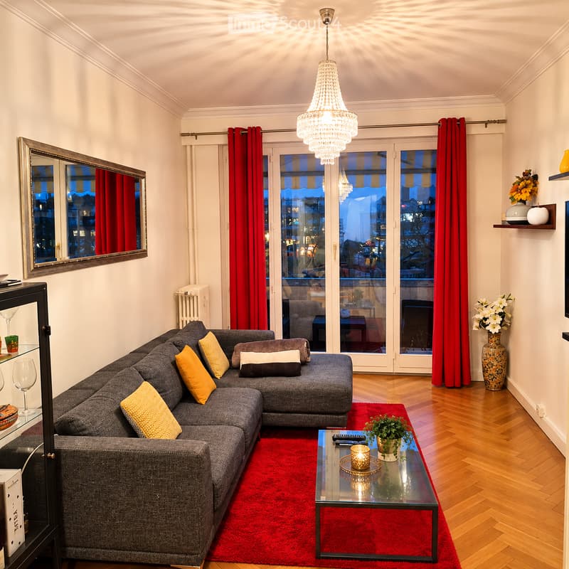 Modern living room with grey sectional sofa, yellow throw pillows, glass coffee table, red area rug, large mirror, decorative vases, and a chandelier.
